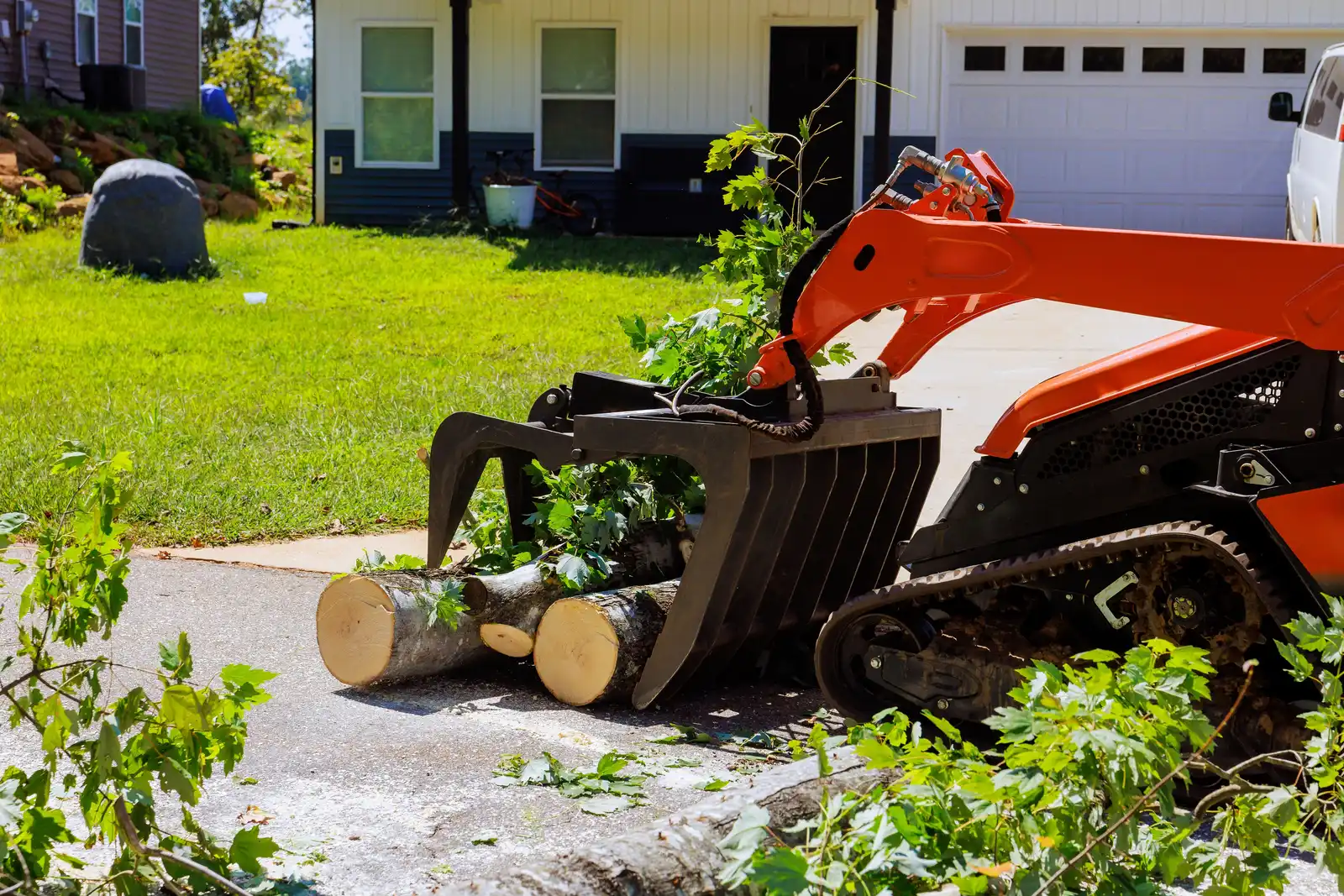 An orange tracked mini loader with a grapple attachment holding cut trunk sections on a residential driveway